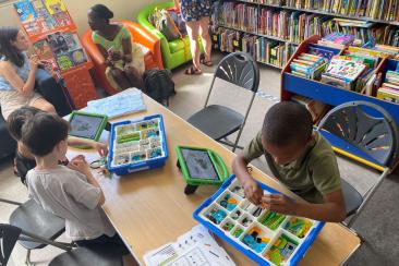Children sat at a table doing different activities in a library, there are bookshelves and two parents talking in the background