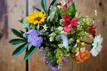 a bouquet of flowers photographed from above, they are sat on a wooden table. There are green fronds, daisies and purple, yellow and orange larger flowers.