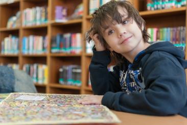 Colour photo of young child reading a book with library shelves in the background