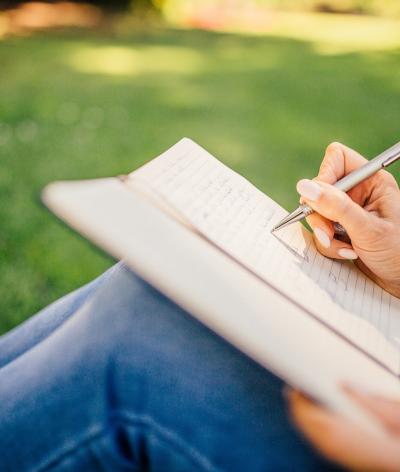 Colour photo of person sitting in a park writing in a notebook