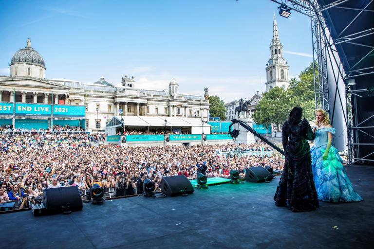 The witches Elphaba in a black dress and green makeup and Glinda in blue dress with blonde hair singing to a crowd in Trafalgar Square