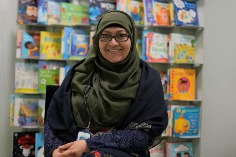 Mahbuba is wearing a headscarf and is seated, smiling and looking directly at the camera, in front of a wall of colourful children's books