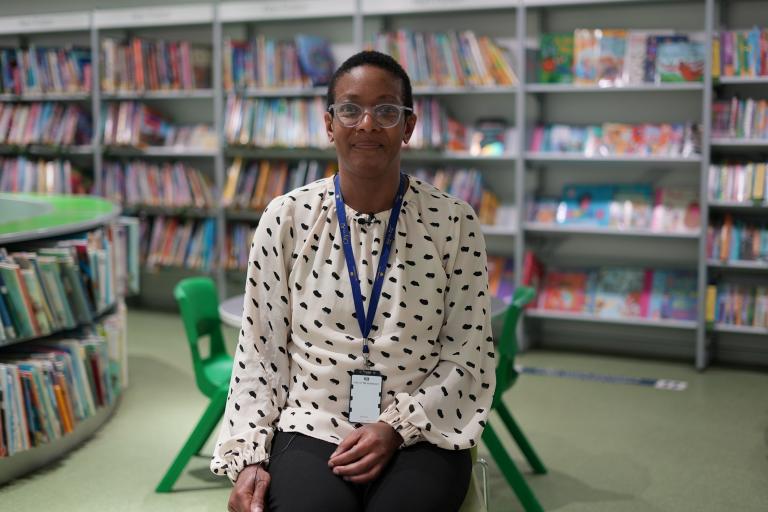 A black, woman librarian sat on a chair in front of a large selection of children's books