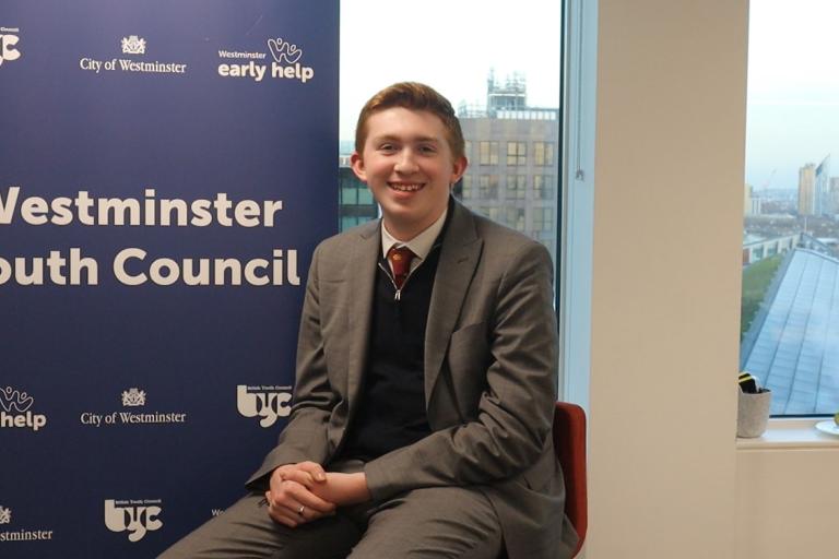 James is seated and smiling at the camera, he is sat next to a blue banner that says 'Westminster Youth Council'