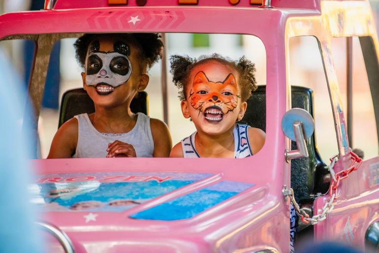 Two smiley girls with their faces painted with animals riding a pink carousel car
