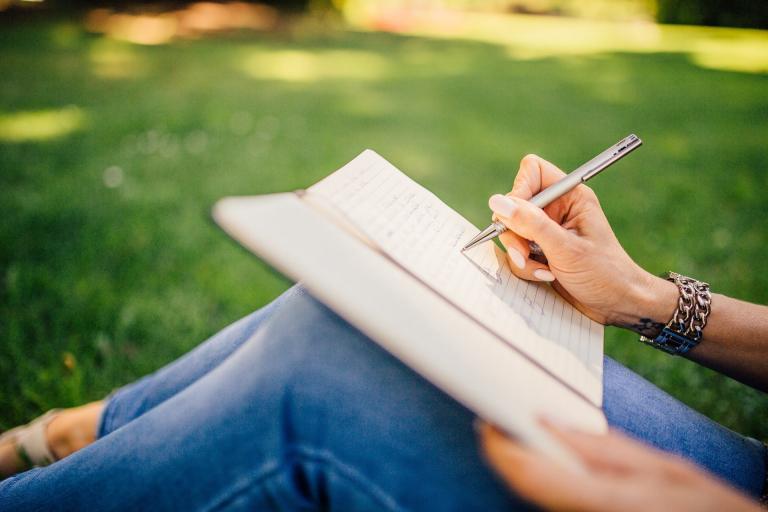 Colour photo of person sitting in a park writing in a notebook