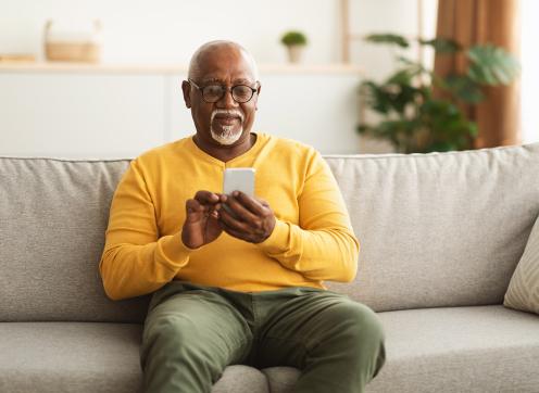 An elderly black male in a yellow sweater using a smartphone while sitting on a sofa