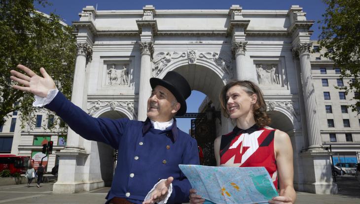 John Nash wearing a dark blue top hat and tails in front of Marble Arch. He is standing with a woman who is holding a map.